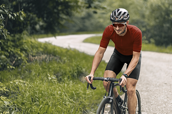 Un hombre vestido con ropa corta de ciclista está sentado en una bicicleta de carreras y pedalea por un camino forestal de grava. Lleva un reloj inteligente Hama en la muñeca.