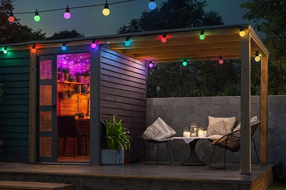 Escena de jardín de noche en verano. Frente a un acogedor salón de madera amueblado, las luces de hadas LED Hama aportan un agradable ambiente al jardín.<br>