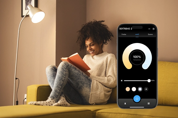 Al fondo, una mujer joven se sienta cómodamente en el sofá bajo una lámpara de lectura con la lámpara LED inteligente Hama WLAN y lee un libro. A la derecha de la imagen se ve un smartphone con la aplicación Smart Home abierta<br>