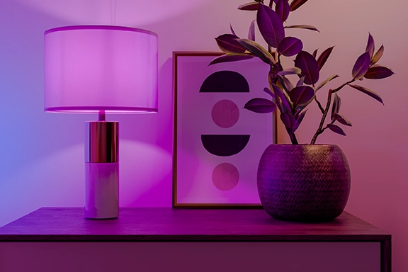 Sobre un aparador de madera hay un jarrón, un marco de fotos y una lámpara que envuelve la elegante lámpara Hama WLAN en una armoniosa luz de color púrpura.<br>