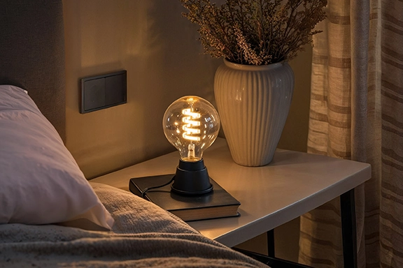 Una lámpara de pie con la bombilla inteligente Hama se alza sobre una mesilla de noche y aporta luz ambiental al dormitorio.<br>