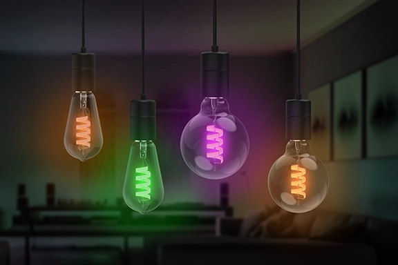 En primer plano se ven varias lámparas Hama que brillan en distintos colores y cuelgan del techo. Al fondo se ve un salón en penumbra.